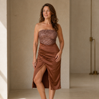 A woman modeling a strapless brown dress with a sequined bodice and a draped satin skirt with a high side slit, standing in a minimalist beige interior.